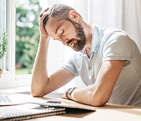 Erschöpfter Mann mit Bart und grauem Polo-Shirt sitzt an einem Schreibtisch und stützt seinen Kopf mit einem Arm